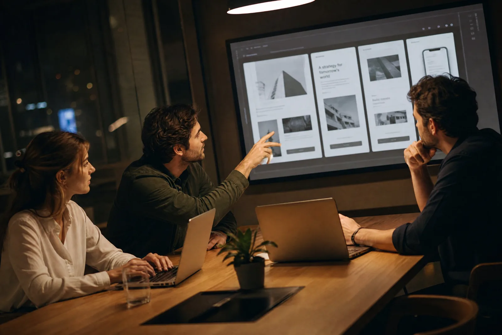 Drei Personen sitzen am Abend in einem Besprechungsraum an einem Tisch mit Laptops, einer zeigt auf einen großen Bildschirm mit Entwürfen einer Website